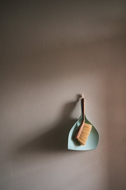 A light blue dustpan and brush hanging on a plain, light-colored wall, with the brush's bristles facing downward. The dustpan is empty and clean, showcasing a smooth surface and simple design. The lig