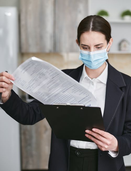 A woman wearing a light blue face mask and formal black blazer with a white shirt is standing indoors in a modern, well-lit room. She is holding a clipboard in one hand and a printed document or clean