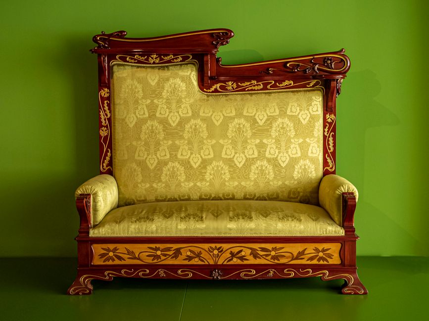 Close-up of an ornate Victorian-style headboard with carved dark wood details and intricate scrollwork at the top. The headboard features a rich, red velvet upholstery with a floral pattern and a velv