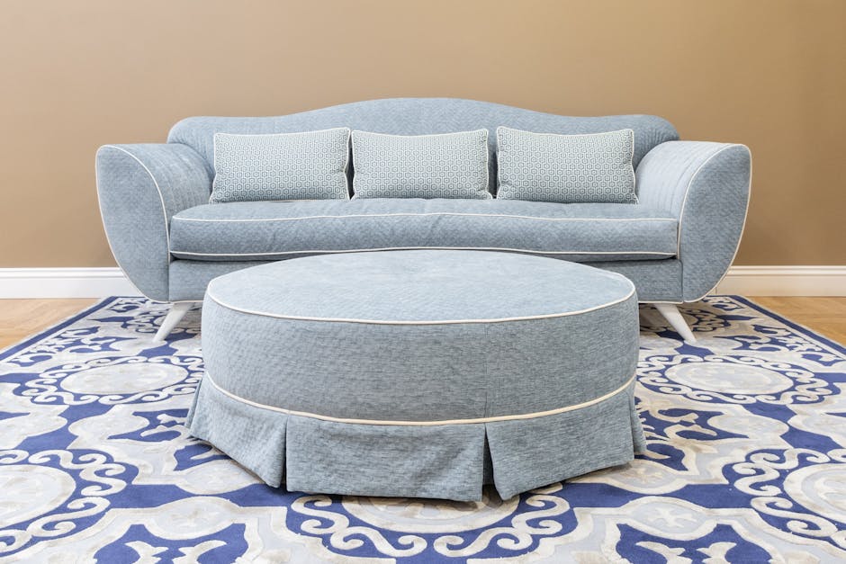 A neatly upholstered light blue sofa with rounded arms and three matching cushions, positioned on a blue and white patterned rug in a living room. In front of the sofa is a round, matching blue ottoma