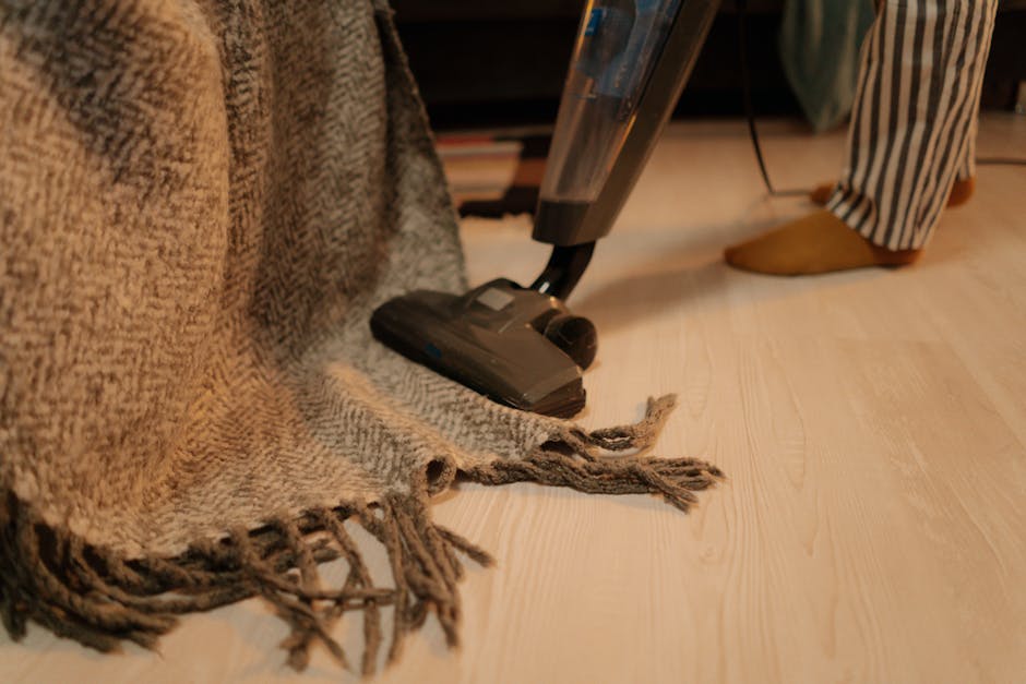 A person kneeling on a patterned carpet in a domestic setting, using a yellow and black vacuum cleaner with a flexible hose and a dustbin to perform surface cleaning. The individual is holding a cloth